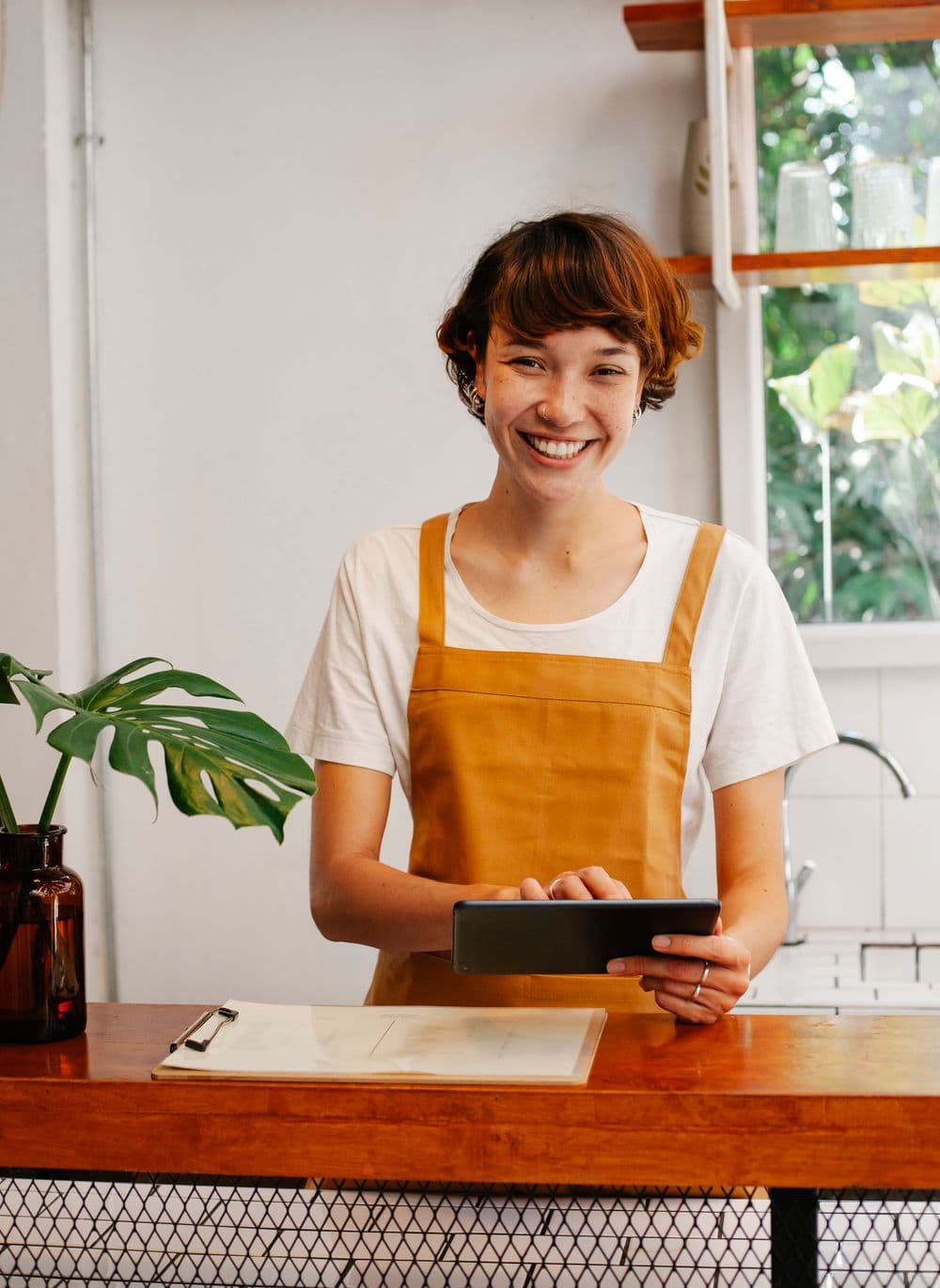 A café owner in an apron smiling behind her counter, working on a tablet.