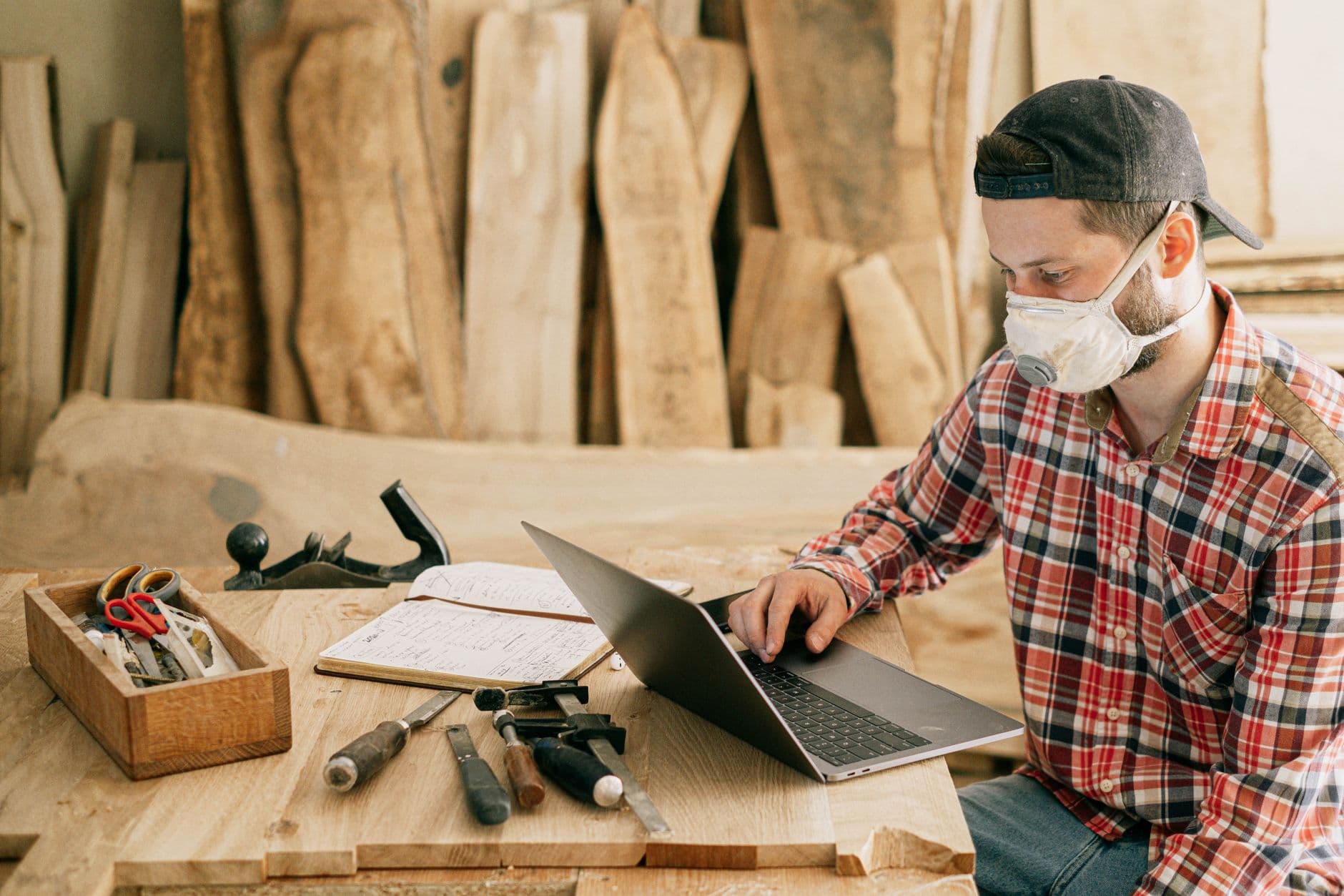 A woodworker at his bench in a workshop, working on a laptop between his tools.
