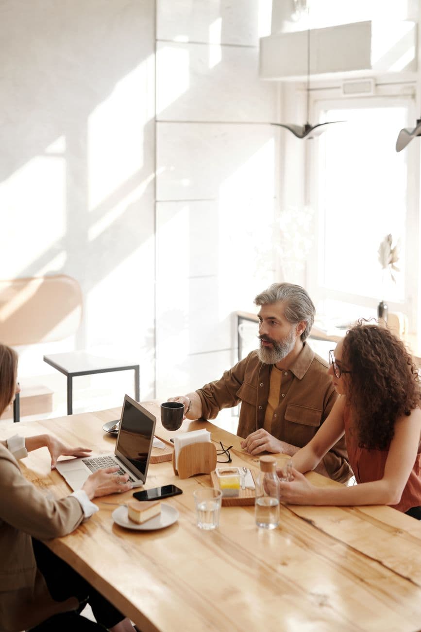 Three colleagues in a coffee shop discussing work at a table with laptops and drinks.