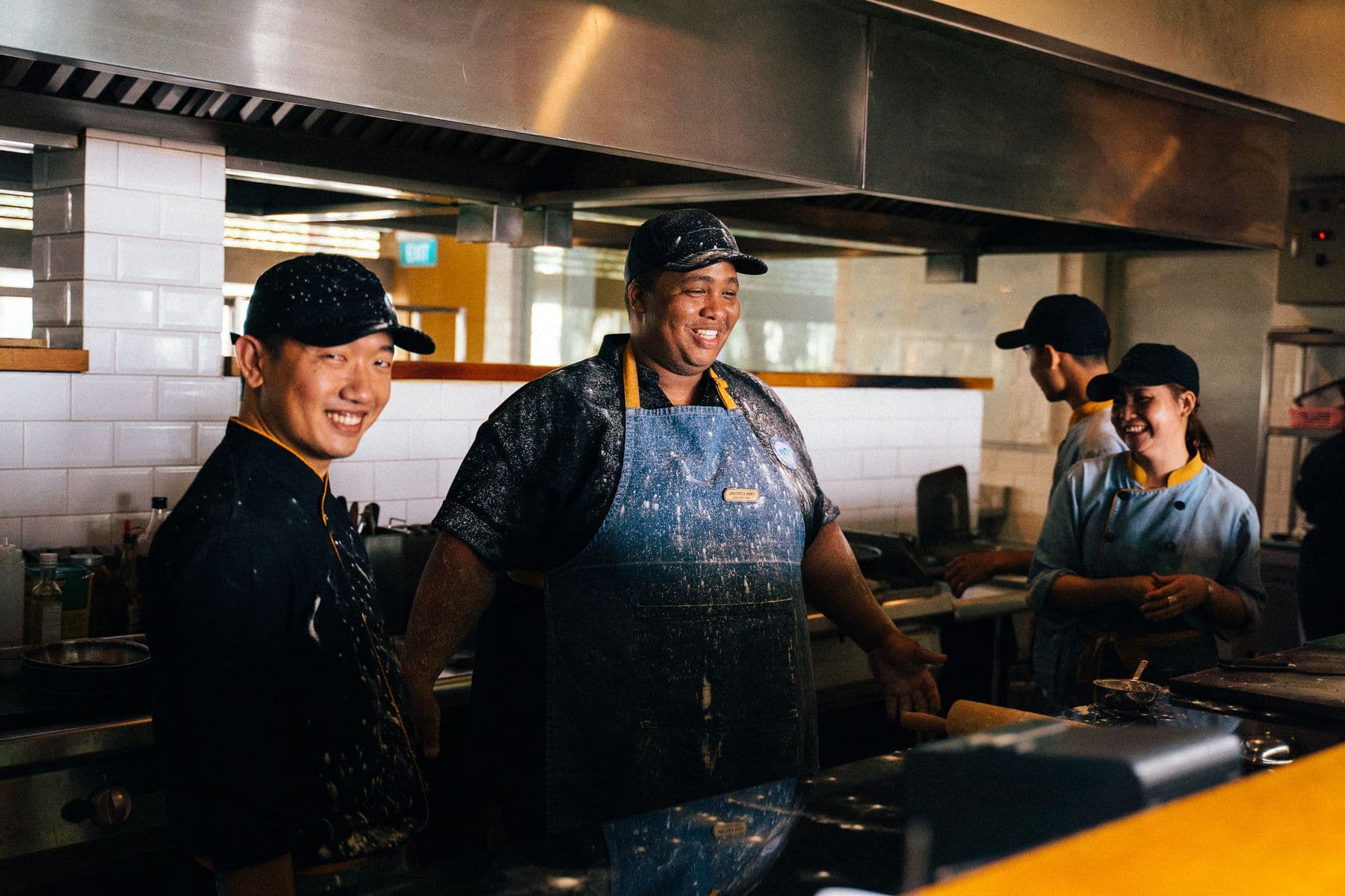 A restaurant kitchen crew in caps and aprons laughing together at the pass.