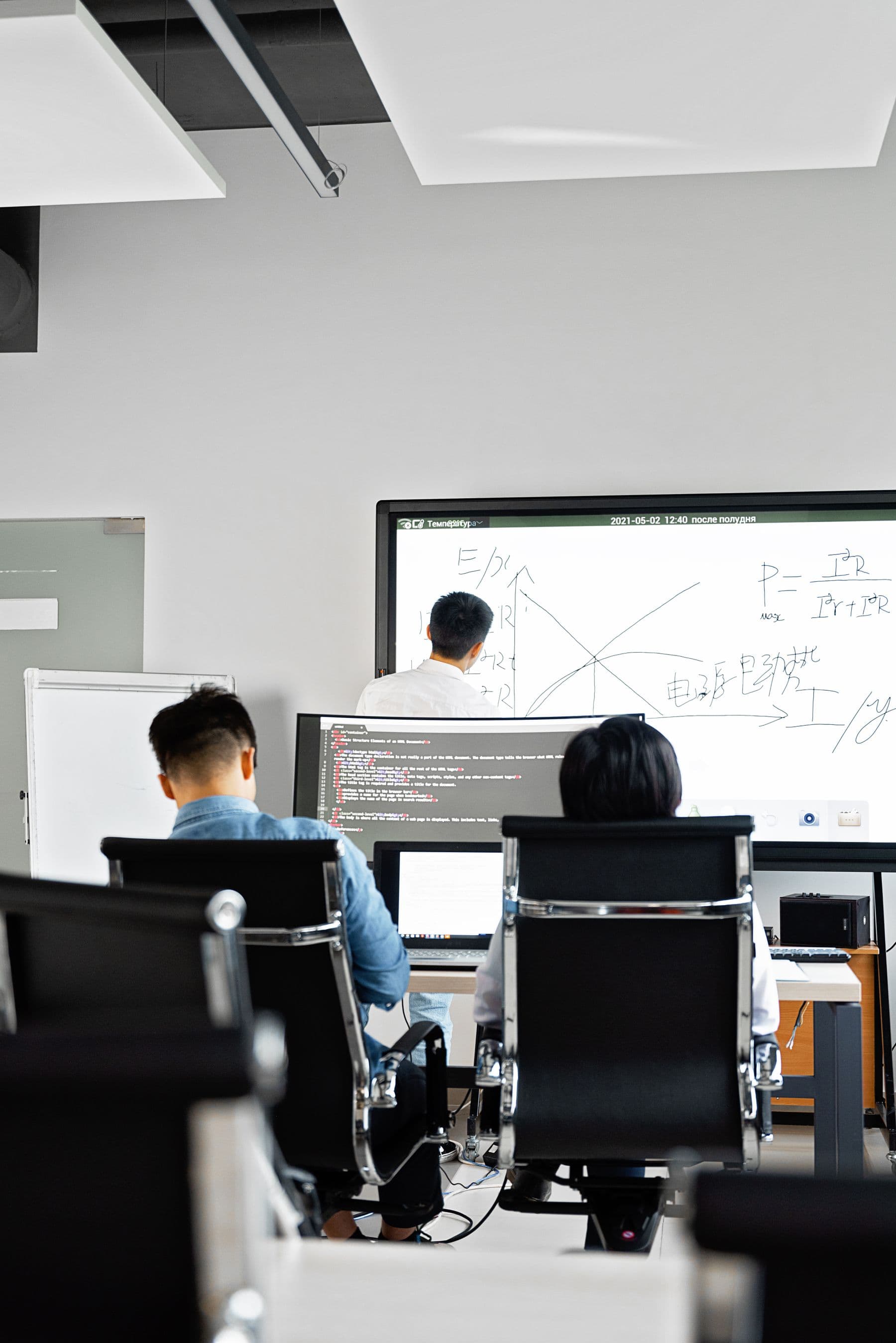 Developers at their screens in an open office, with a large display of charts and code at the front of the room.