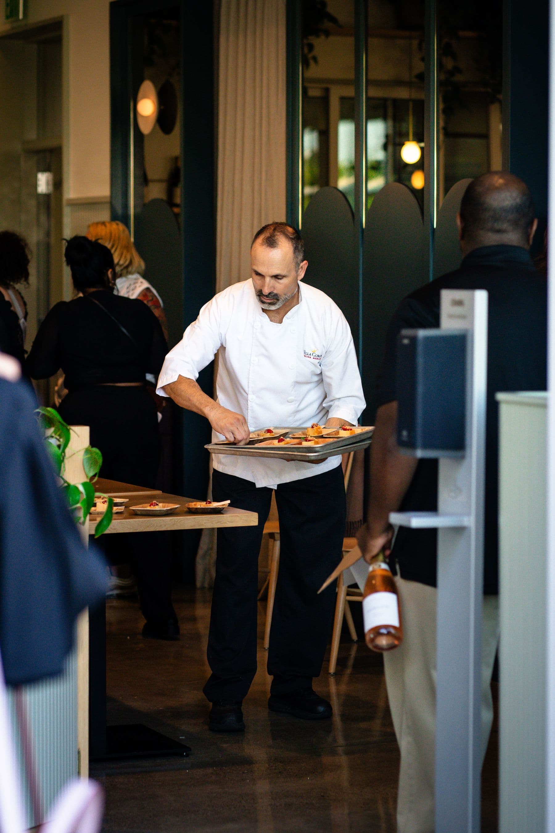 A chef in whites carries a tray of food across a busy restaurant floor during service.