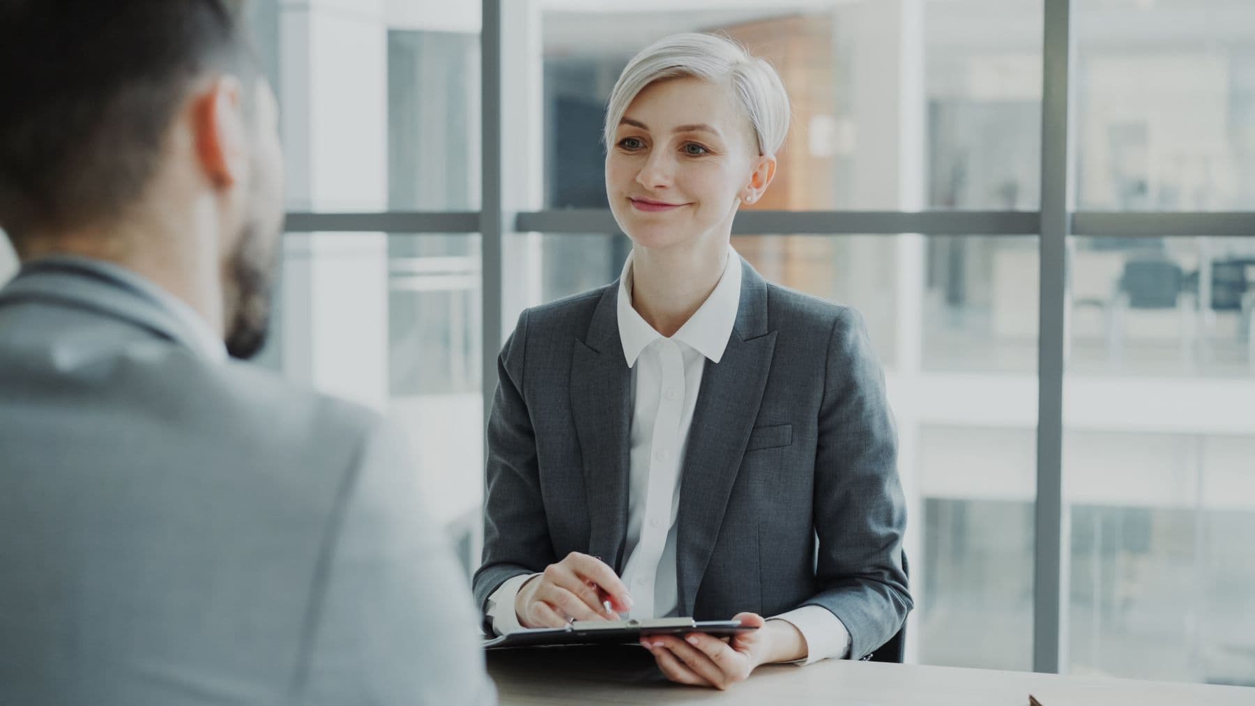 A hiring manager in a grey blazer smiles across a desk at a job candidate in a bright office.