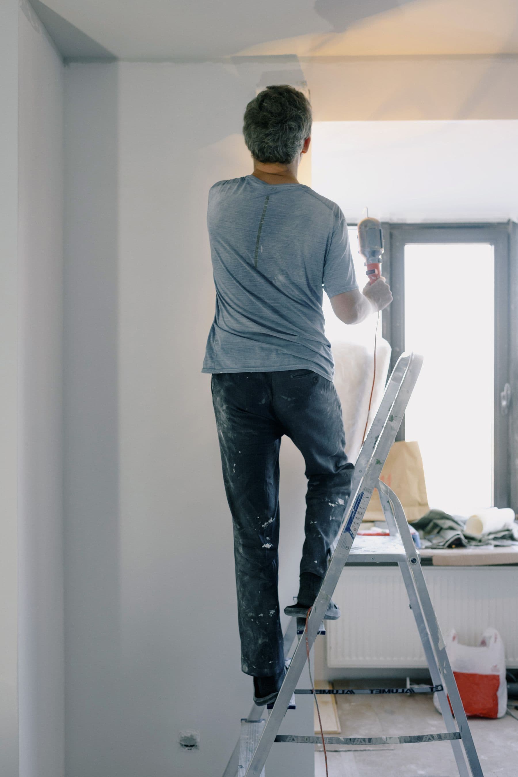 A contractor on a ladder sanding a freshly plastered wall in an apartment under renovation.