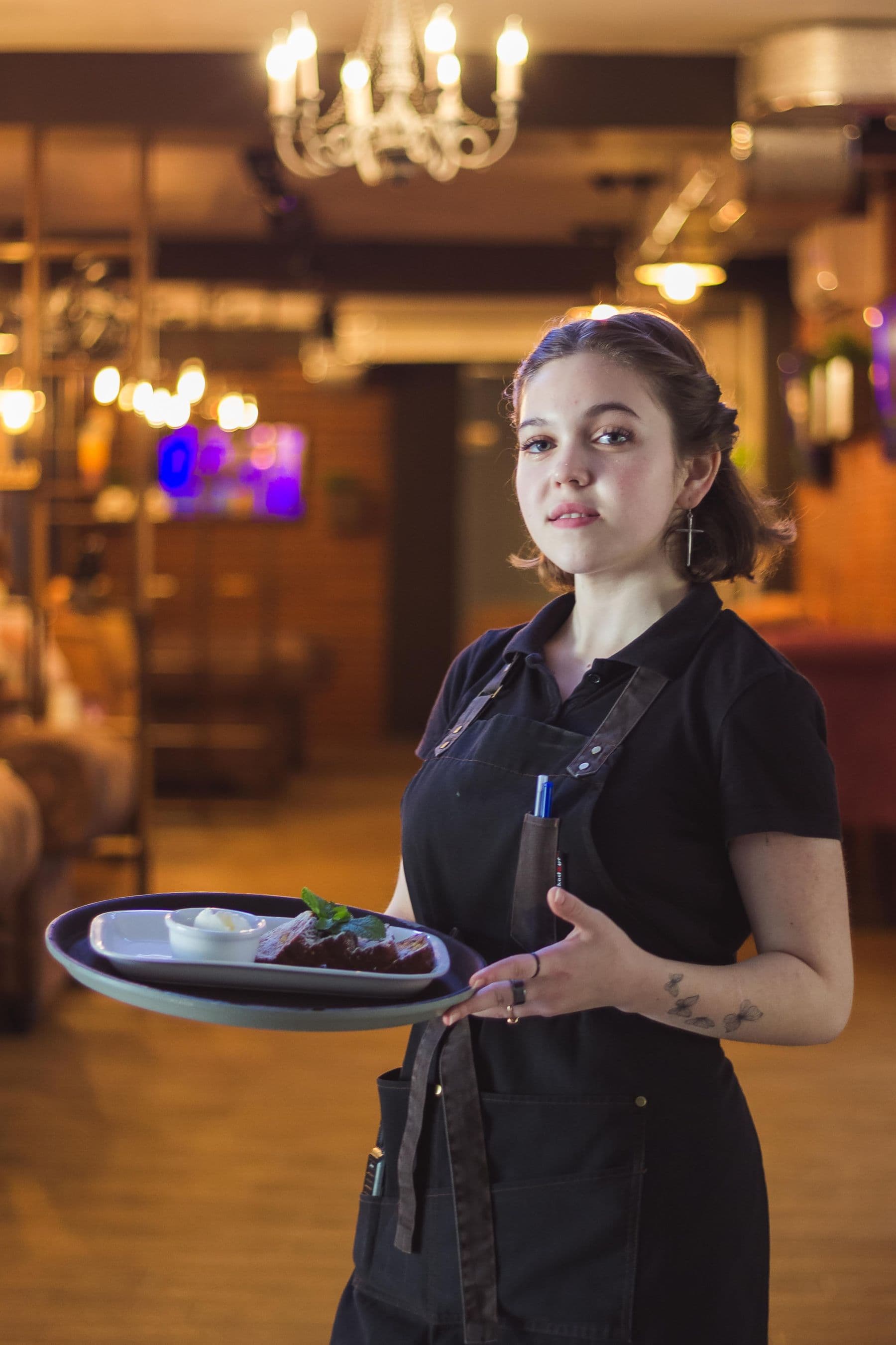 A server in a black apron carries a plated dish through a warmly lit restaurant dining room.